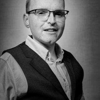 A photograph of Laurence Scott in black and white. He is a short man in his early 30s who wears glasses.